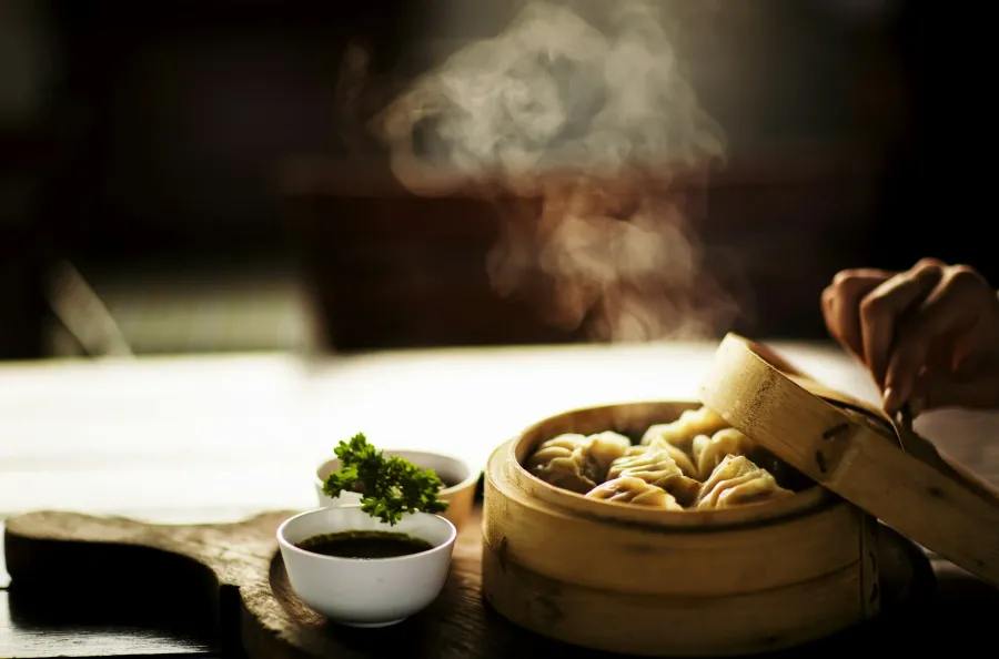 Steaming dumplings in a round wooden basket with dipping sauce